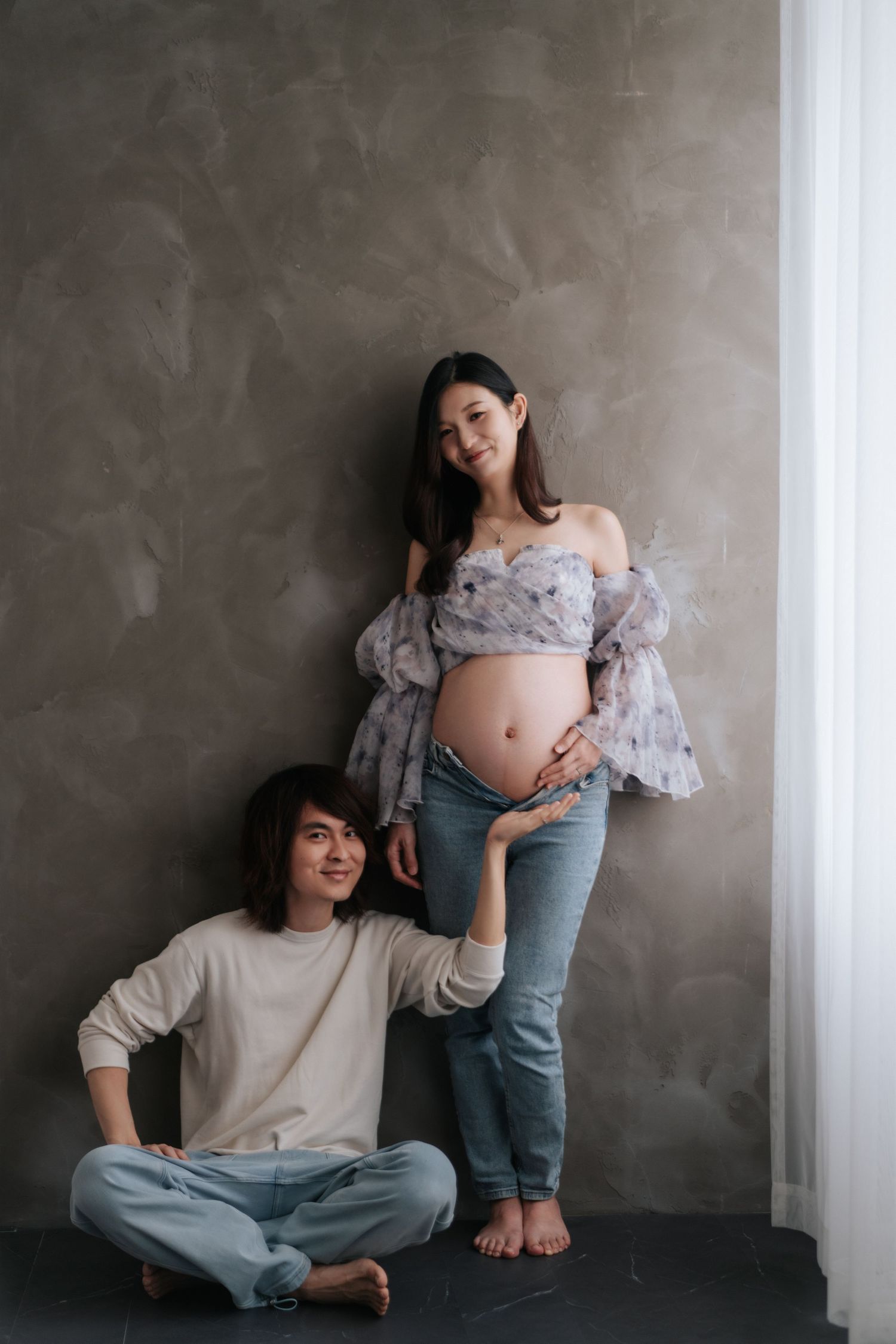 A pregnant woman in casual attire sits against a gray wall while interacting with a small child in denim pants.