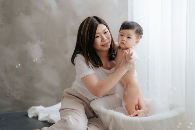 A smiling Asian mother tenderly holds her baby boy while sitting together in a bright, softly lit room.