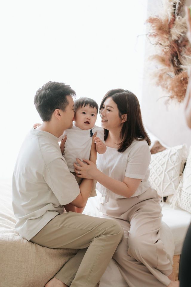 Asian family with parents sitting on bed holding their baby, sharing a joyful moment together in a bright, cozy room.