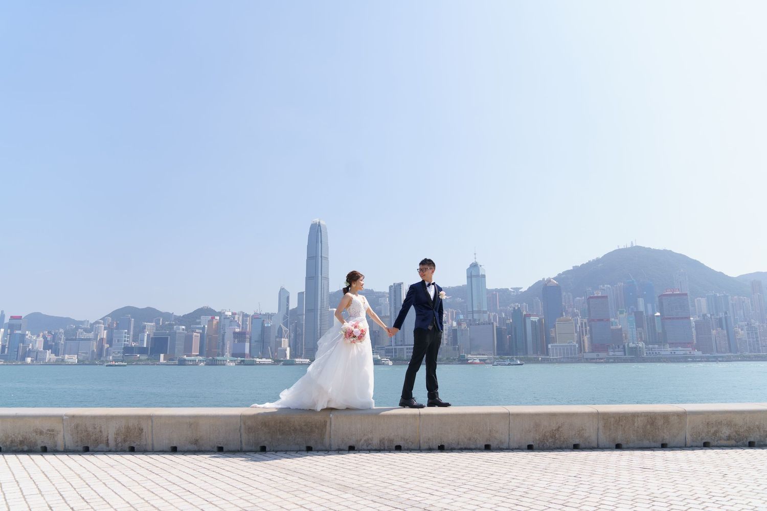 Bride and groom pose on a waterfront promenade with Hong Kong skyline and Victoria Harbour in the background.