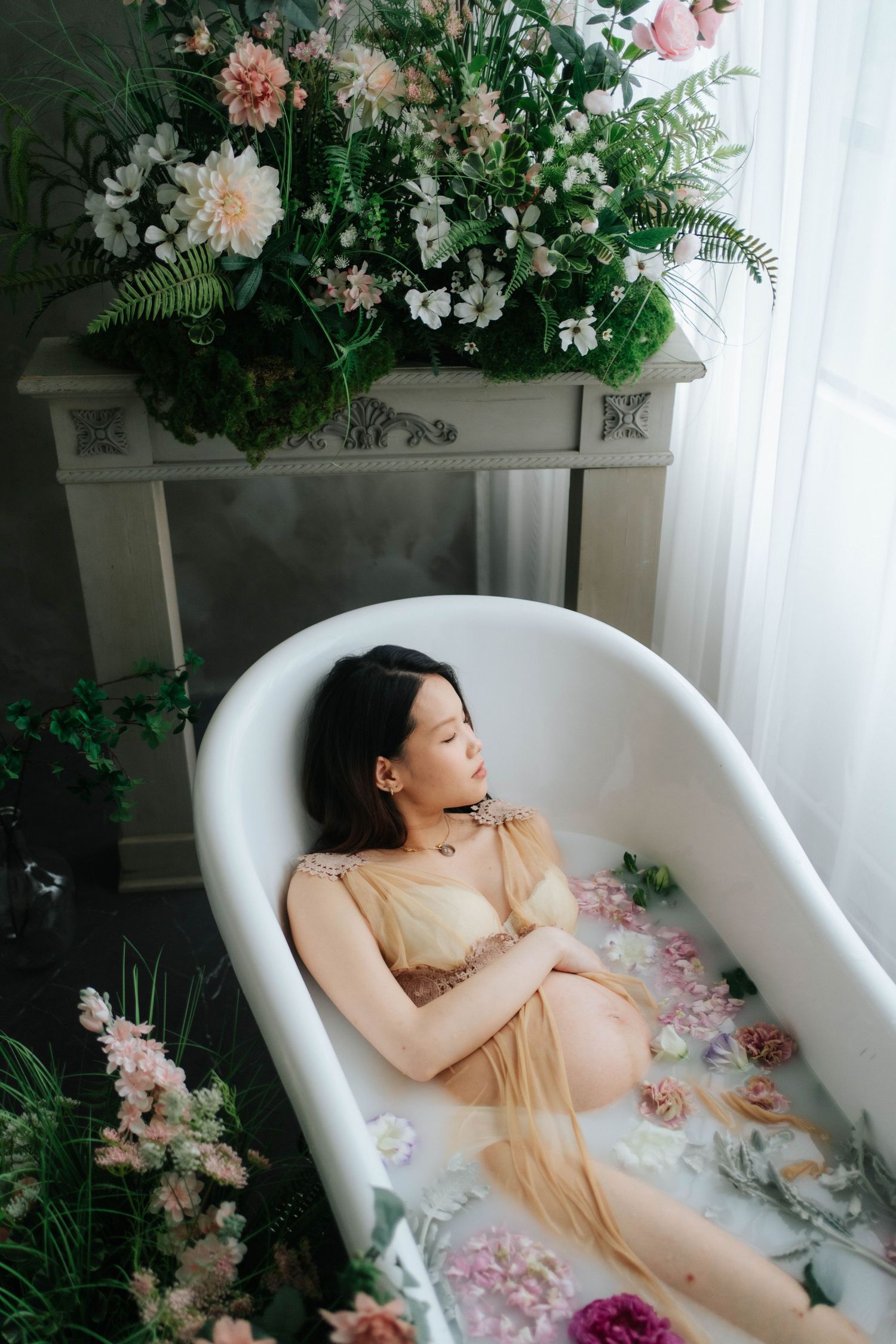 A person relaxes in a white bathtub surrounded by pink and white flowers and greenery in an ethereal setting.