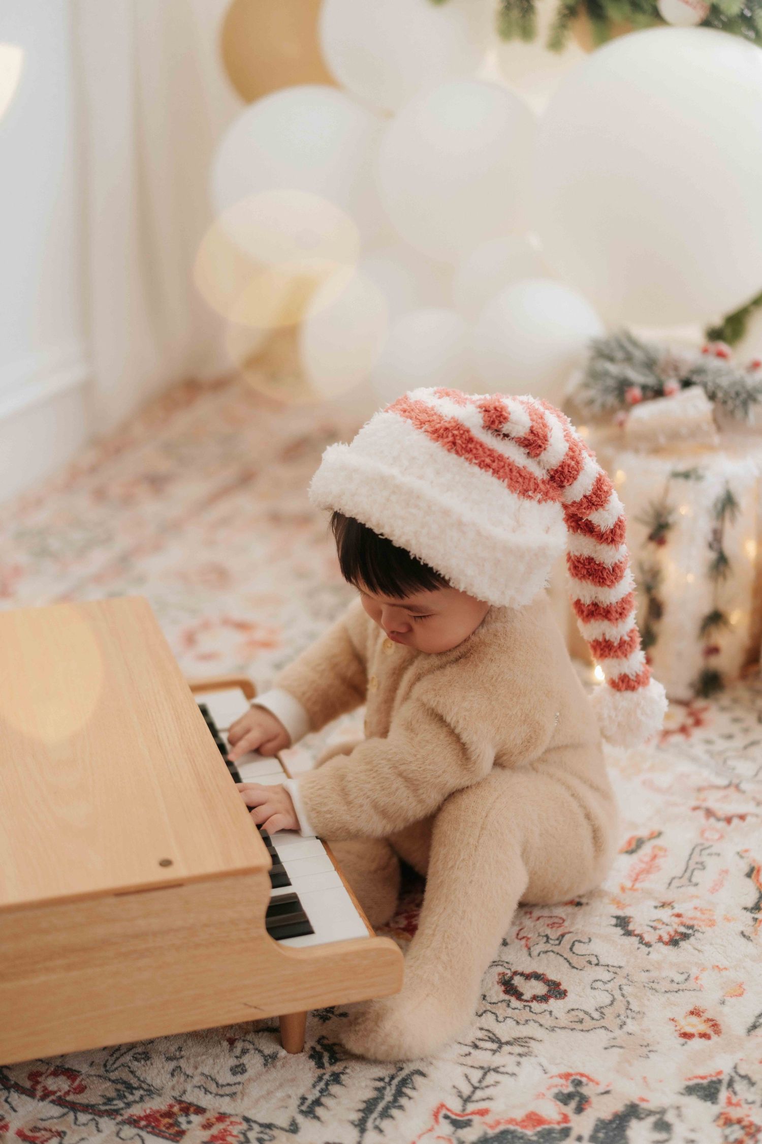 Toddler in fuzzy bear outfit and striped Santa hat playing toy piano on rug with balloons and Christmas tree.