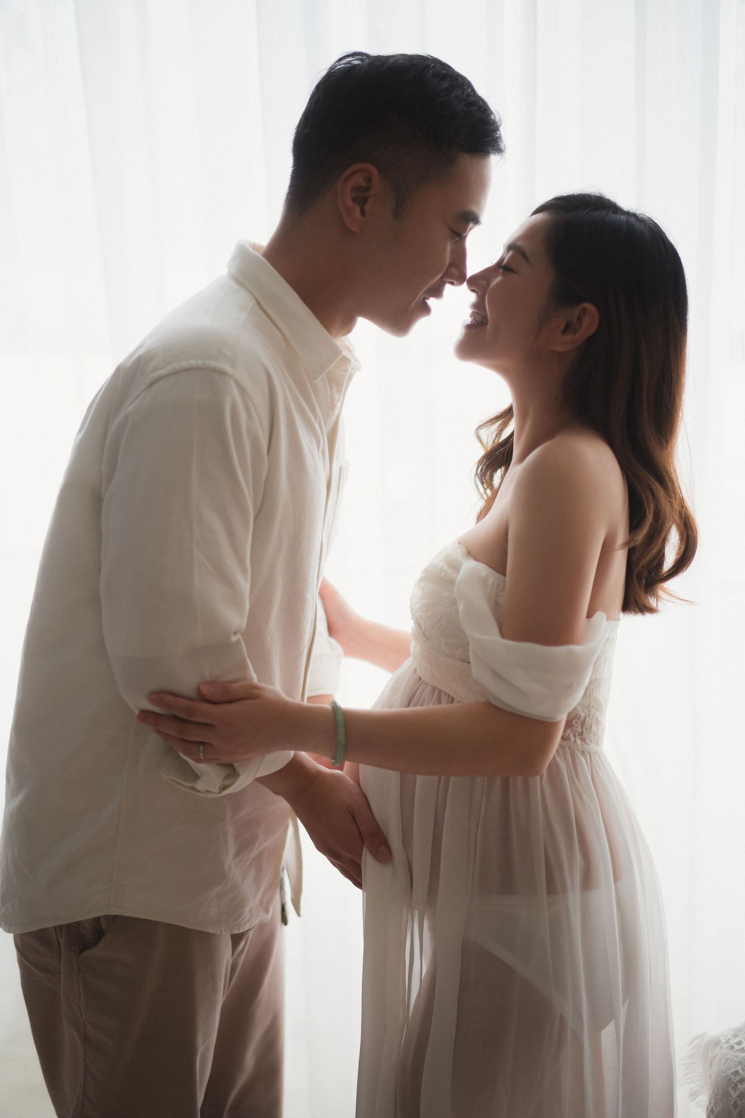 A romantic maternity photoshoot shows tender moments between a couple in white outfits against a light background一群浪漫的孕婦寫真，展現了一對身著白色服裝的夫婦在淺色