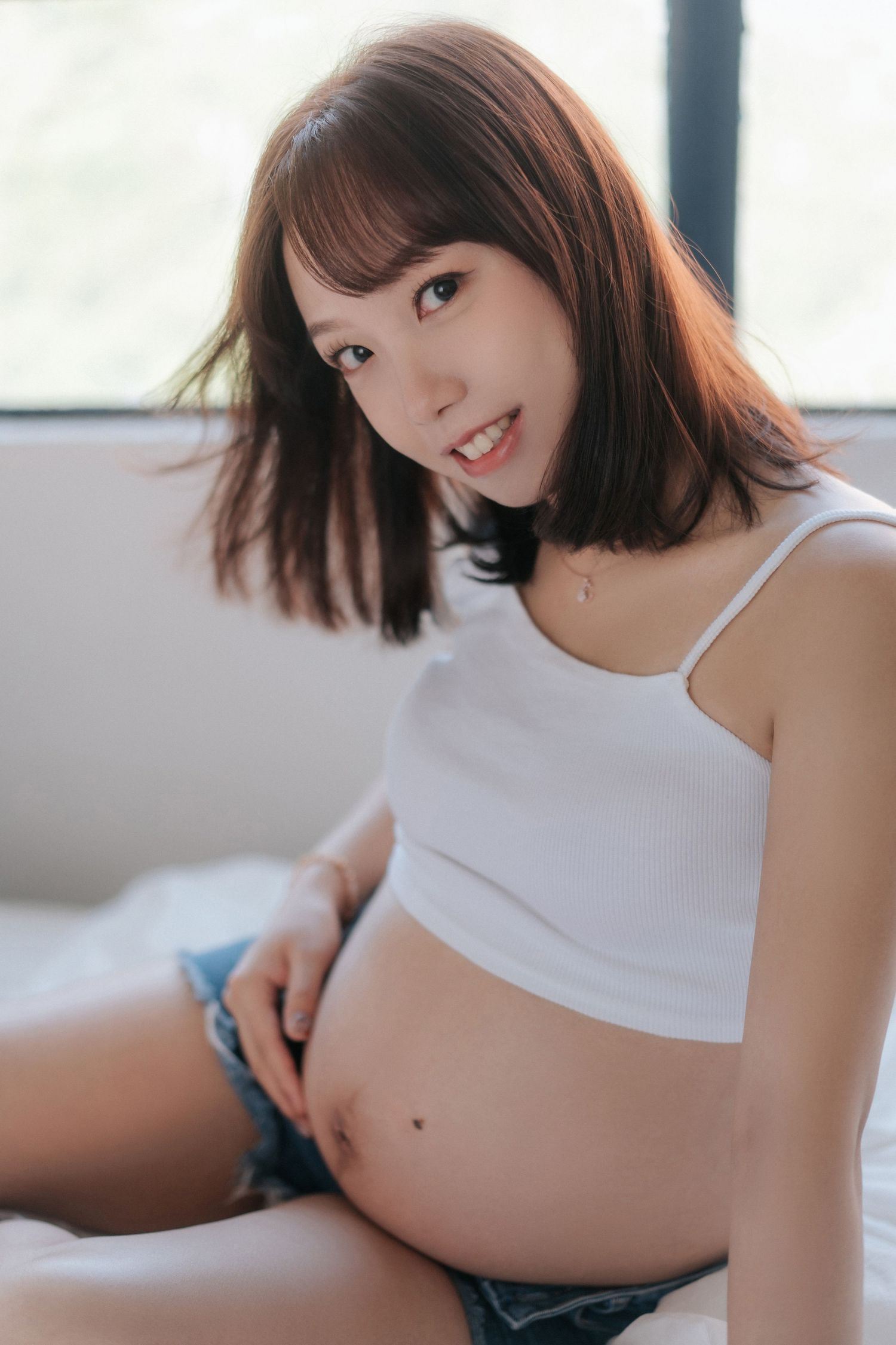 Young woman in white crop top and denim shorts sits casually by window in natural lighting.一位身穿白色露臍上衣和牛仔短褲的年輕女子隨意地坐在窗邊，沐浴著自然光。