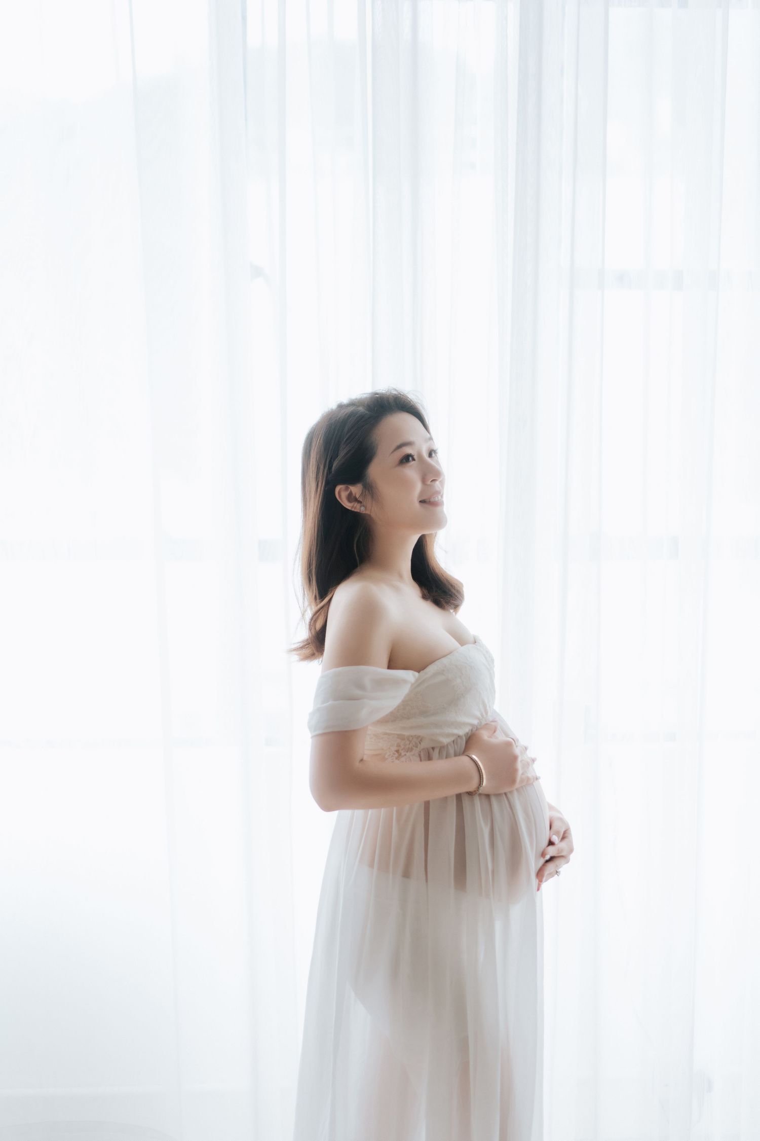 Expectant mother in flowing white maternity dress poses gracefully against bright window backdrop in studio setting.