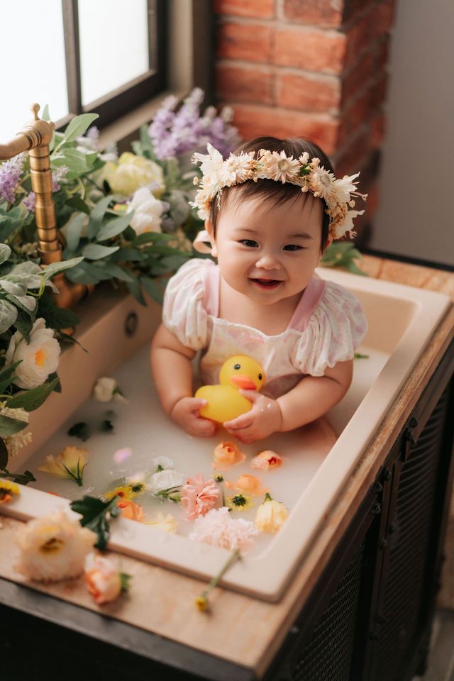 Smiling baby girl in floral crown holds rubber duck in milk bath surrounded by colorful flowers near window.