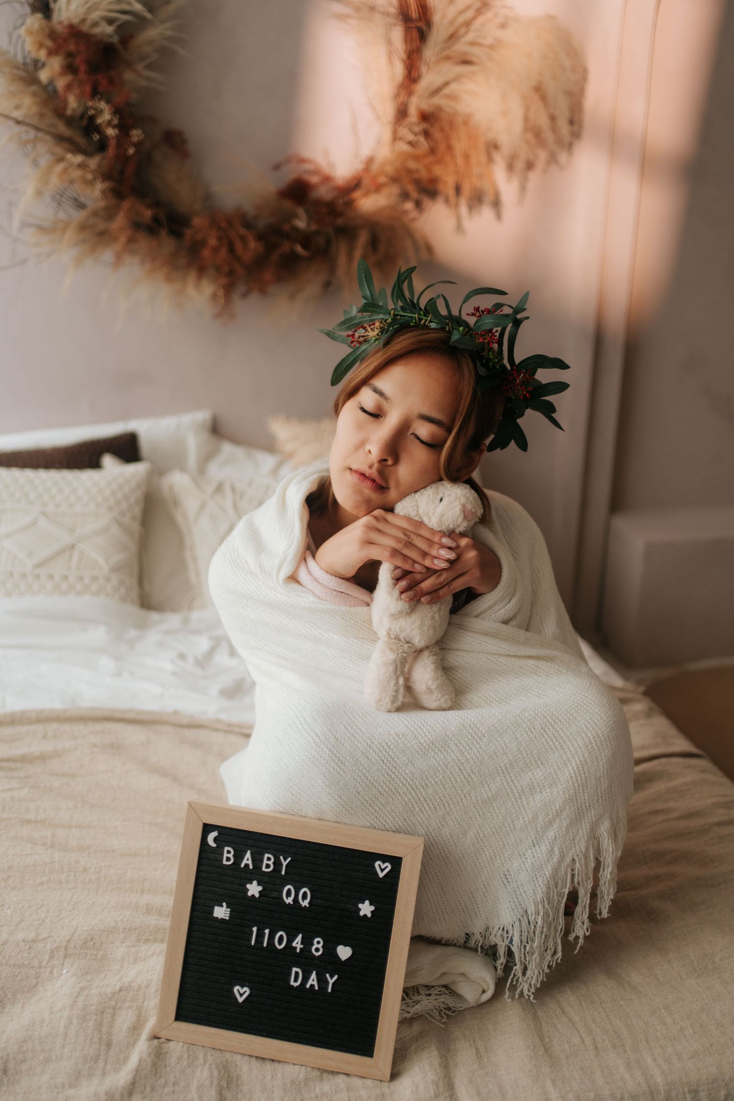 Pregnant woman in floral crown holding stuffed bunny on bed beside a letter board reading Baby QQ 11048 Day.