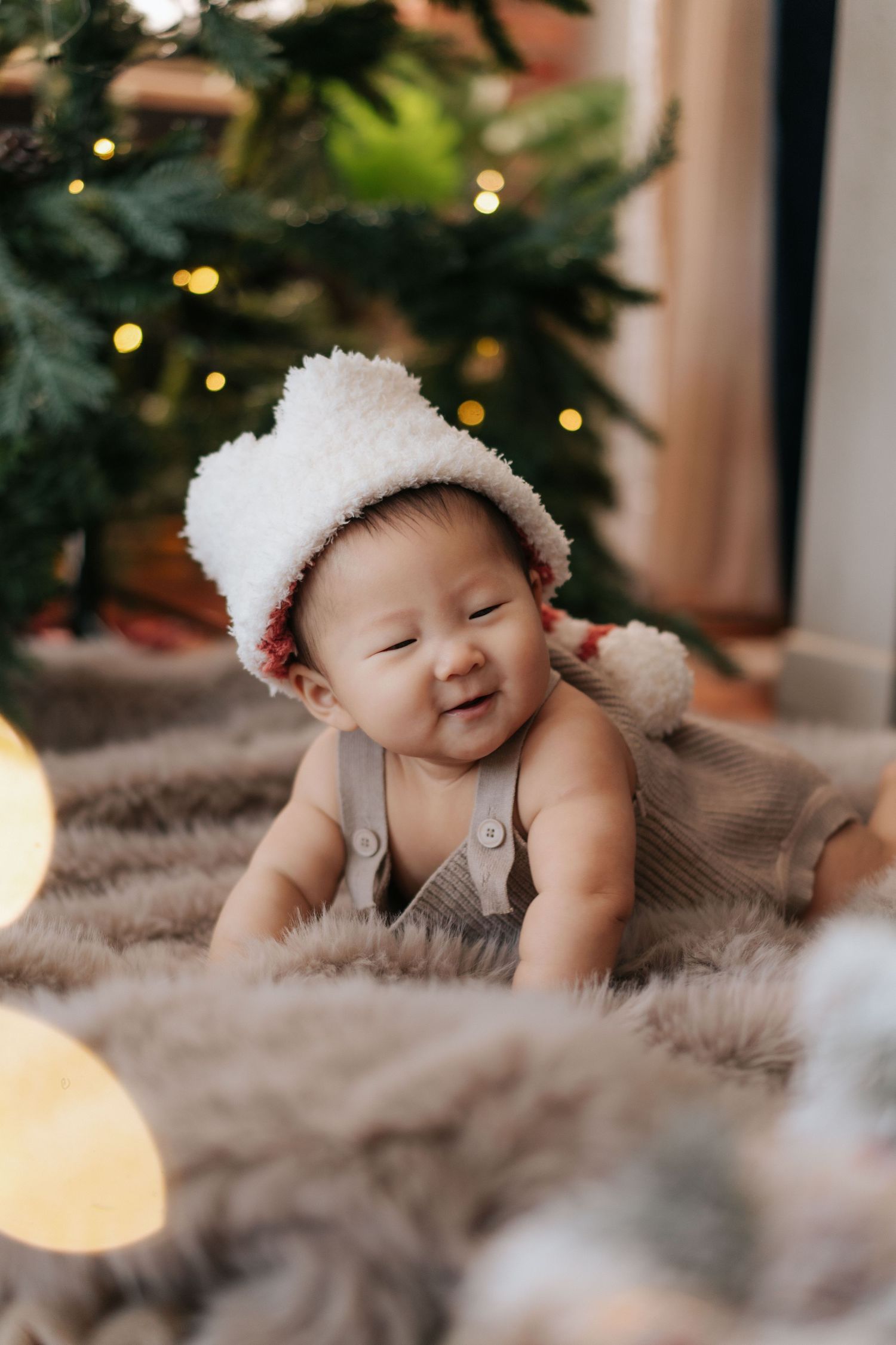 Adorable Asian baby in white fluffy hat and overalls smiling on fur rug near Christmas tree with lights.
