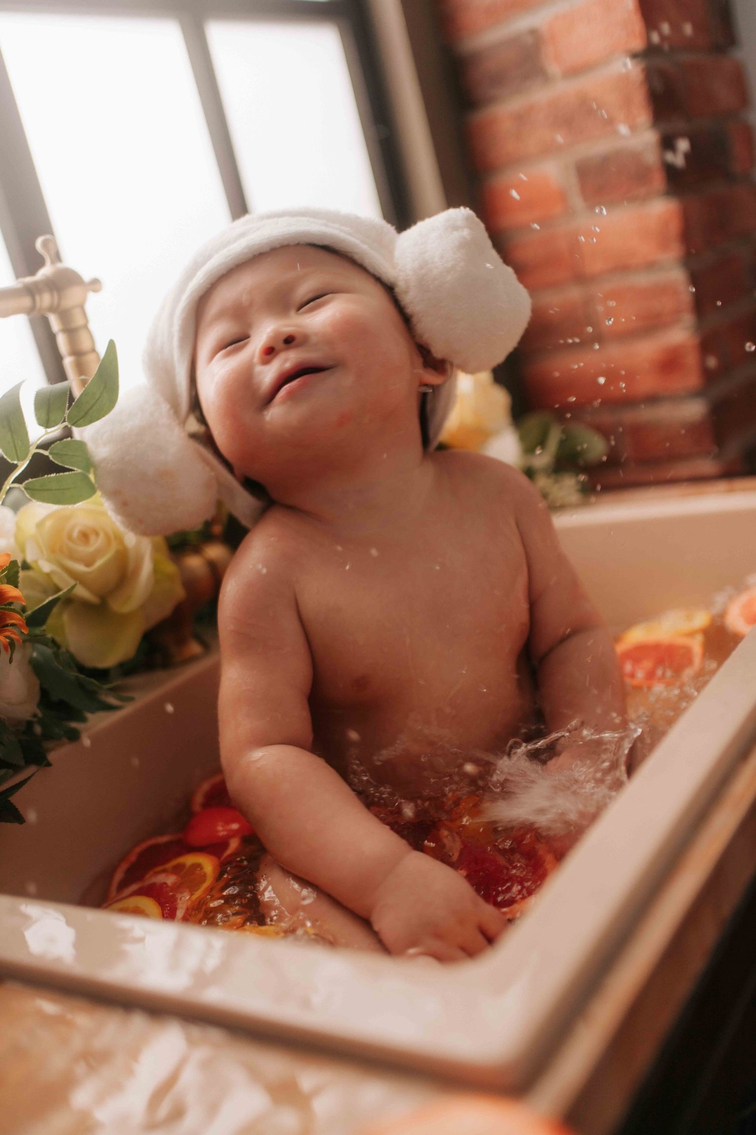 A baby wearing a white hat lies in a bathtub surrounded by yellow roses and red flower petals.一個戴著白色帽子的嬰兒躺在浴缸裡，周圍環繞著黃色玫瑰和紅色花瓣。