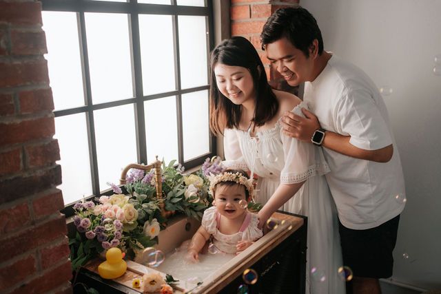 Happy Asian family with parents smiling over baby in floral bath setup near window with brick wall.
