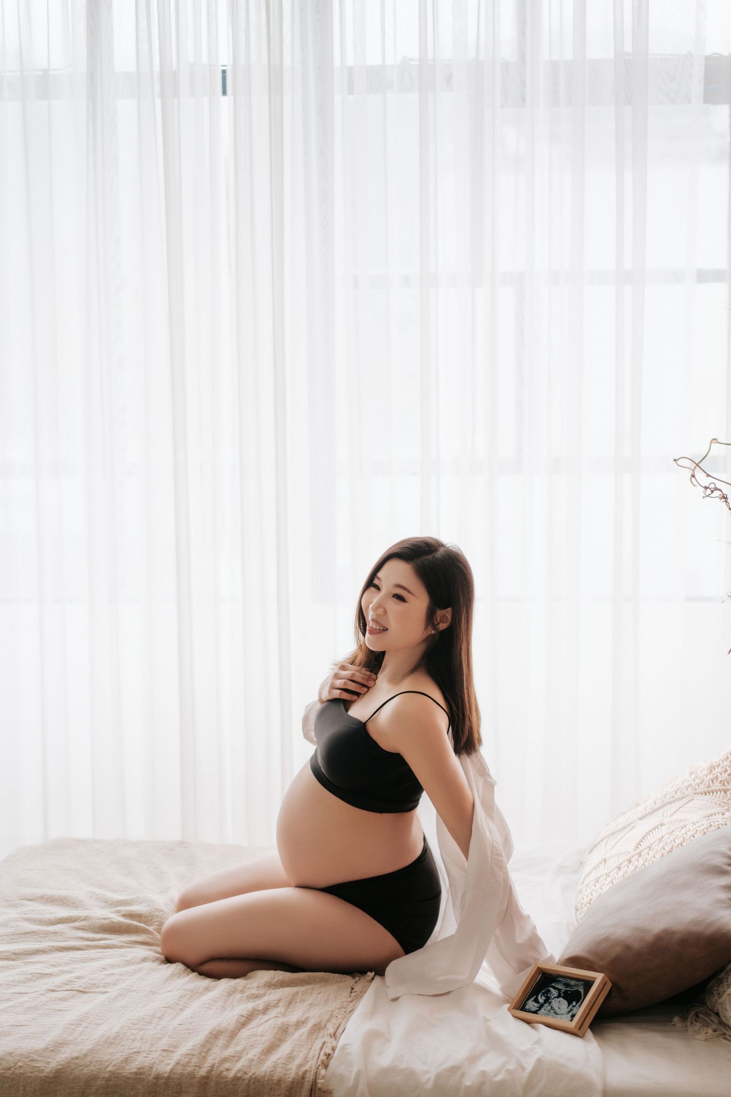 A pregnant woman in black loungewear relaxes on white bedding in a bright, airy bedroom setting.