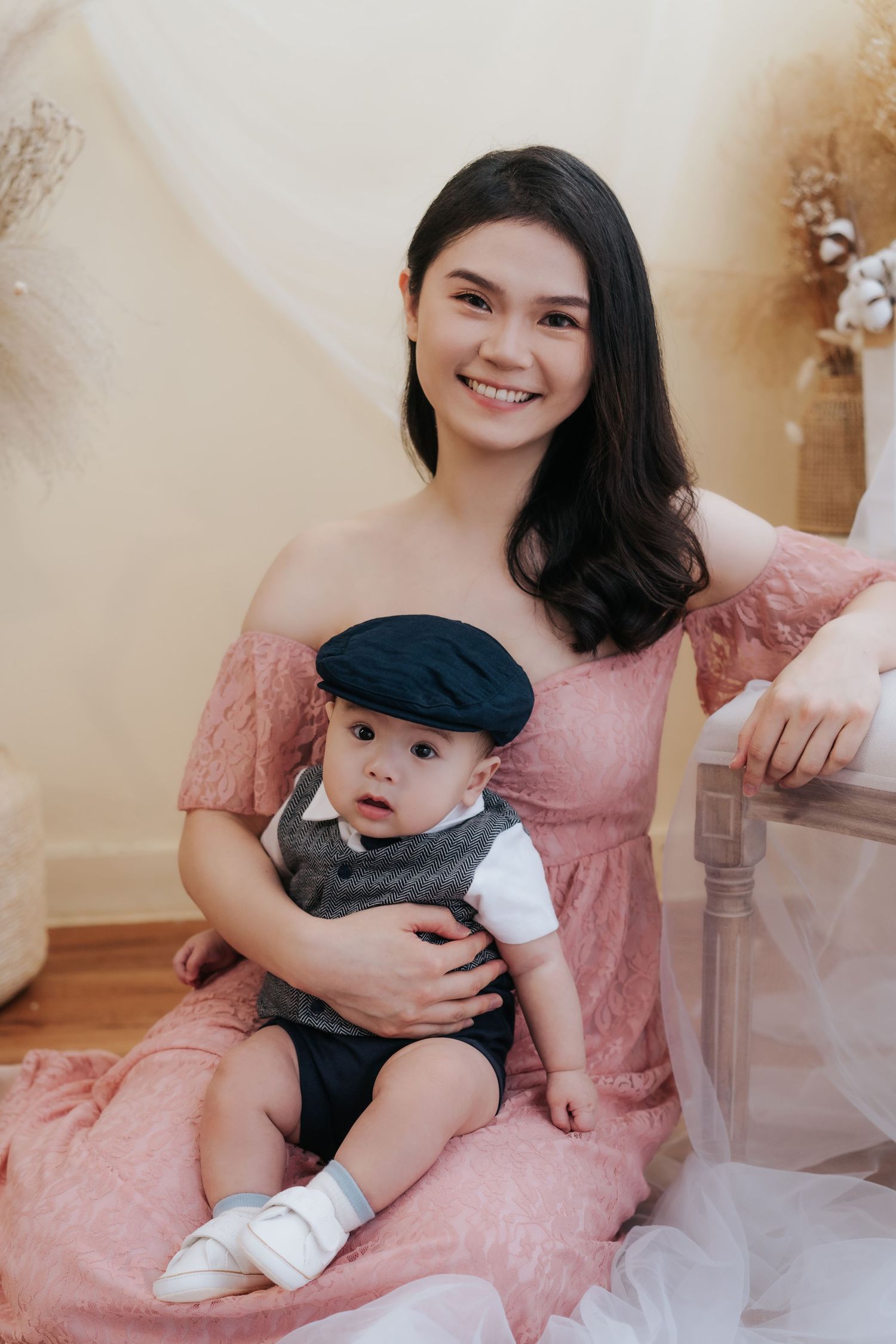 A mother in a pink dress holds her baby wearing a black cap in a tender indoor photoshoot series.