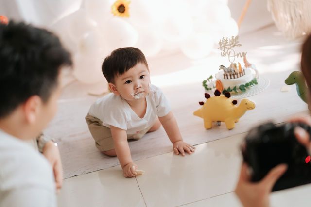 A baby crawls on the floor near a dinosaur toy and birthday cake during a first birthday photoshoot.