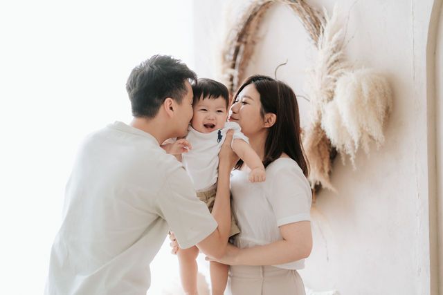 Happy Asian family with parents kissing their smiling baby in a bright, minimalist white studio setting.