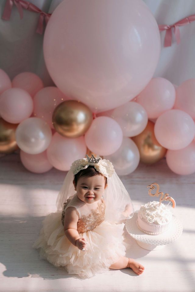 Smiling baby girl in white tutu dress and crown sits beside first birthday cake with pink and gold balloon backdrop.