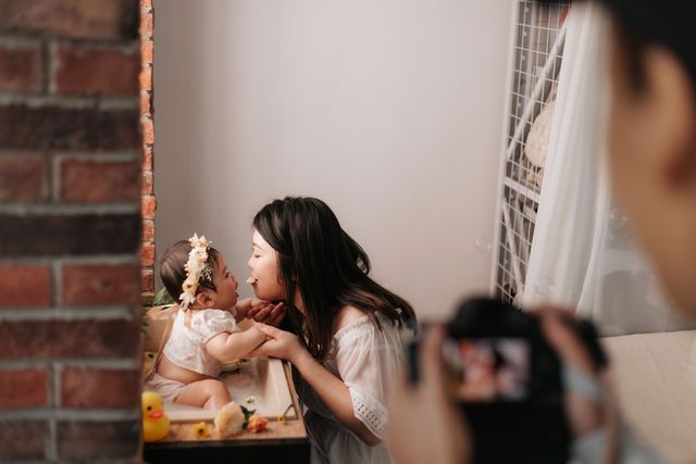 Mother in white dress tenderly interacting with baby on decorated table while photographer captures the moment.