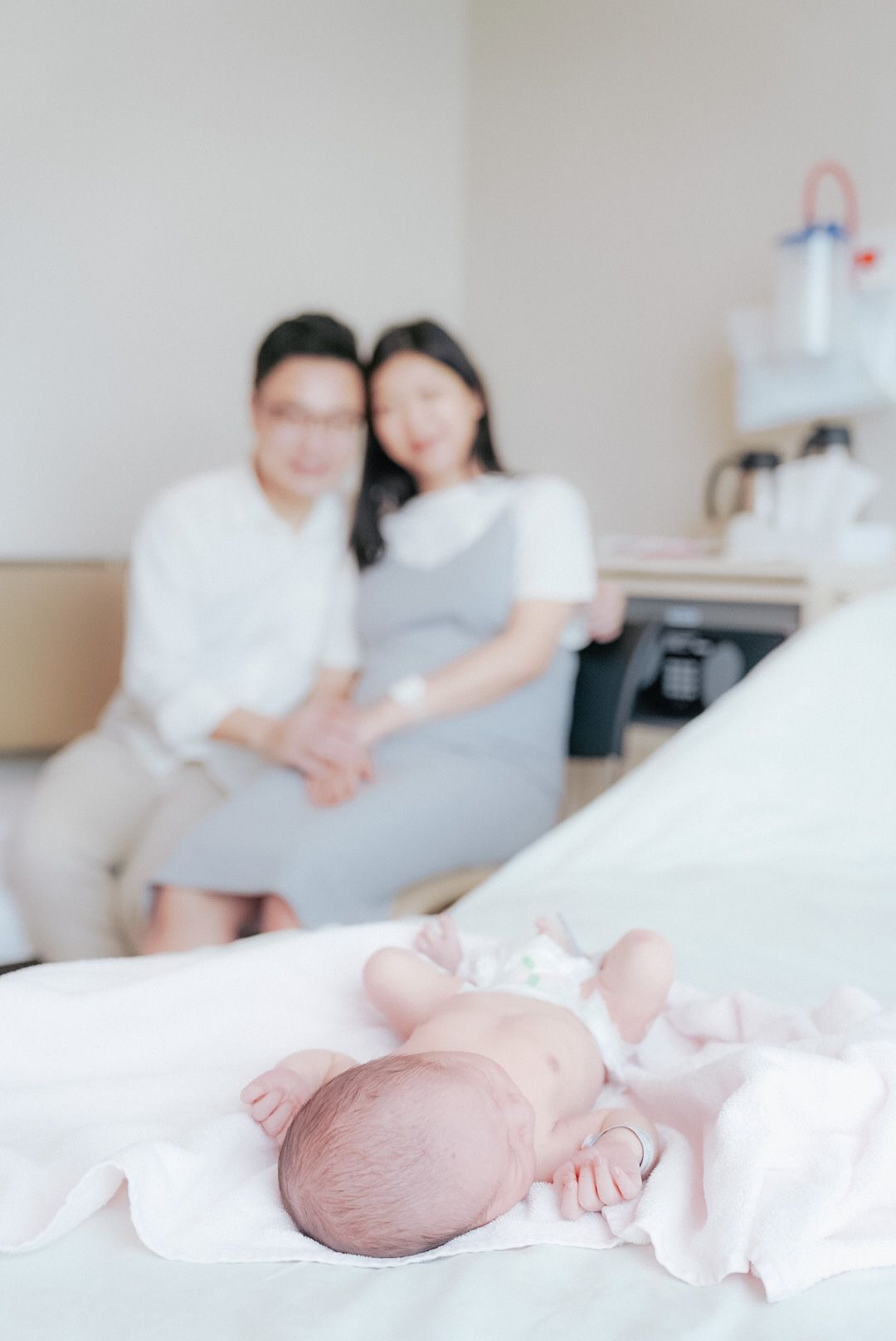 A newborn baby lies peacefully on white bedding while parents sit nearby in a bright, minimalist setting.新生兒安靜地躺在白色的床單上，父母坐在附近，環境明亮簡約。