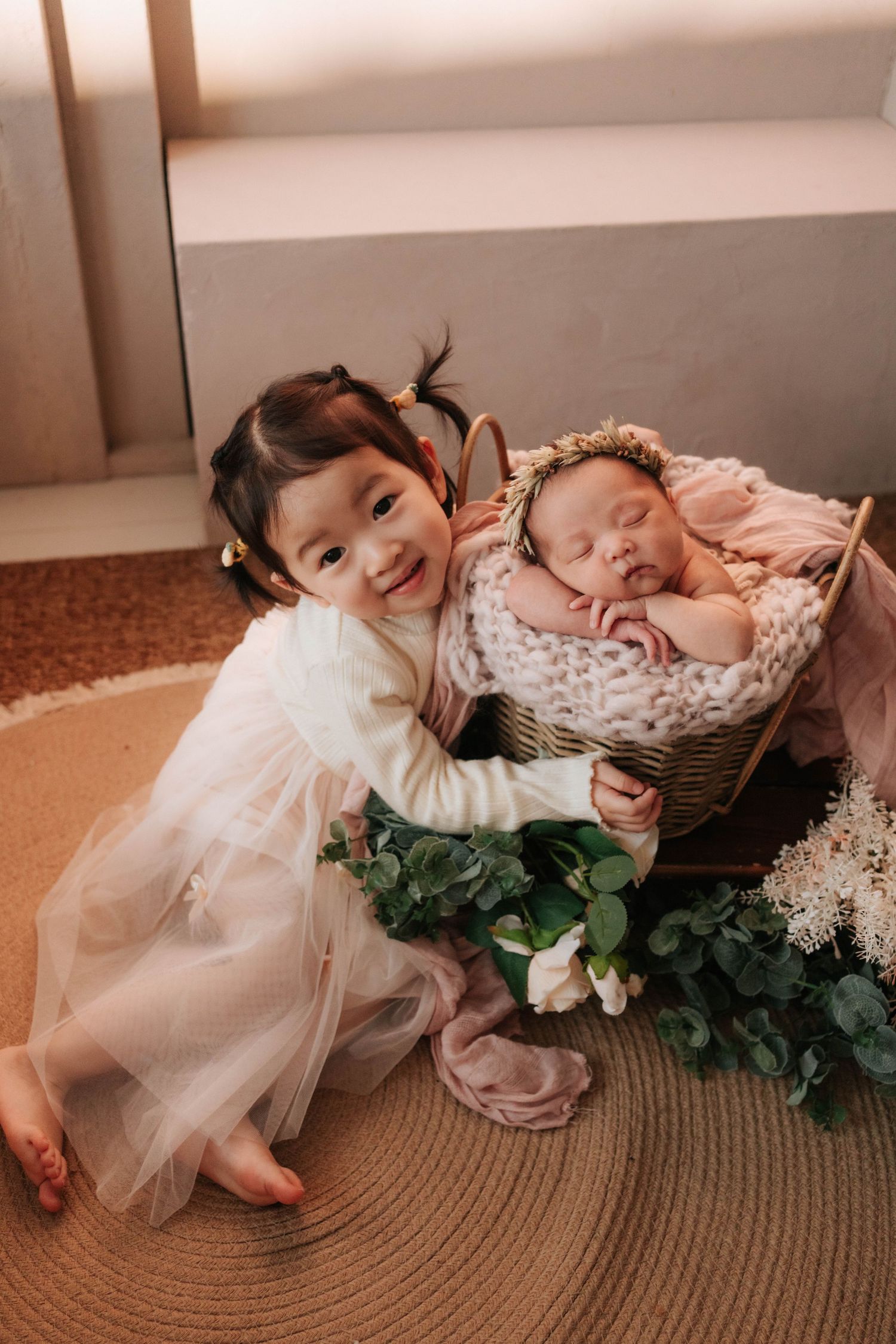 A young child in a white dress sits beside a wicker basket with a sleeping baby, surrounded by greenery and flowers.