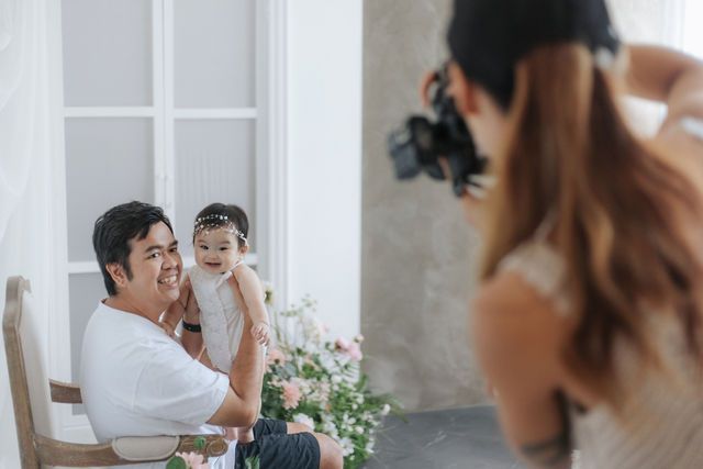 Family photoshoot with father holding baby girl while photographer takes pictures in bright studio.