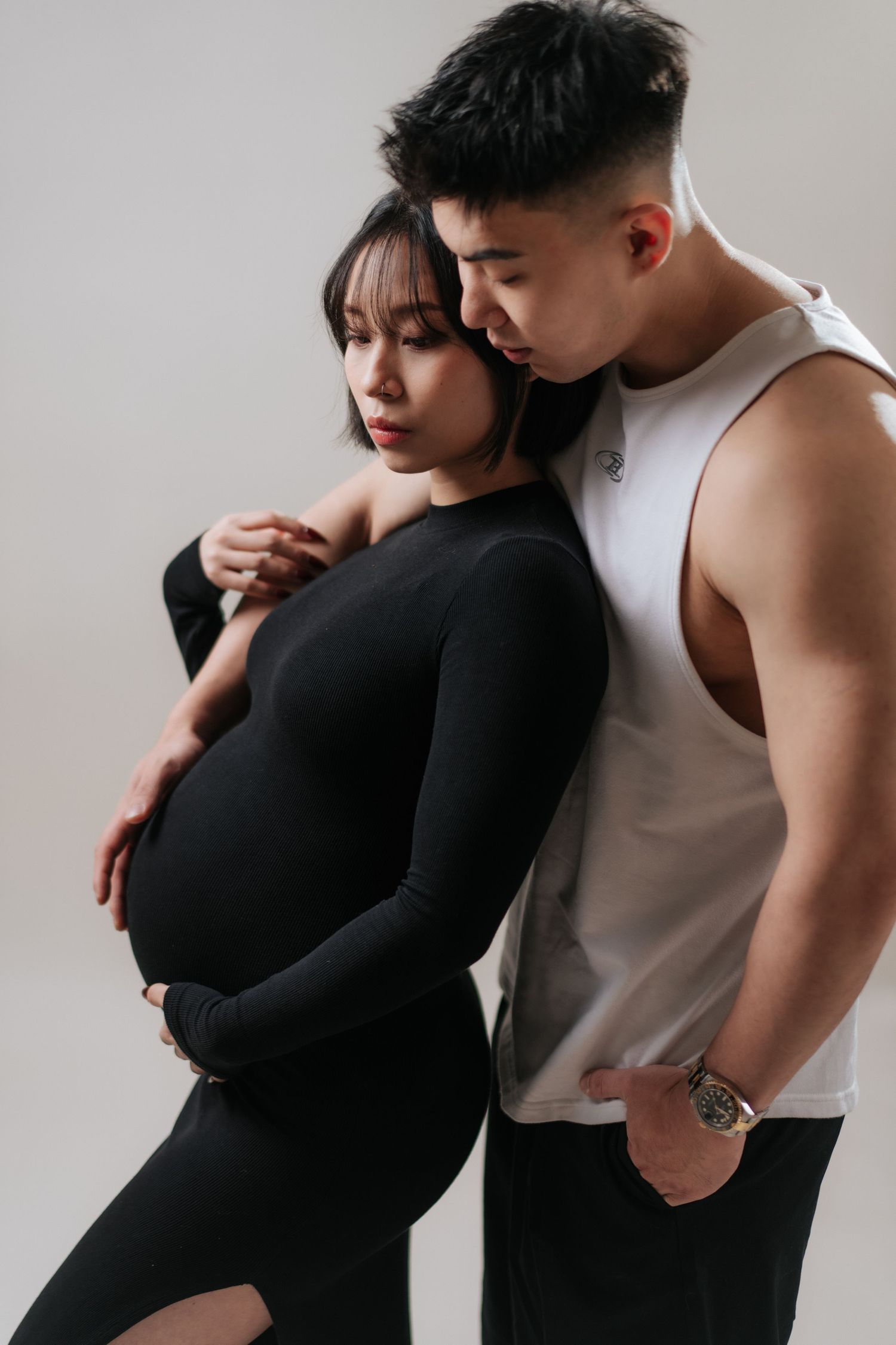 A pregnant woman in a black dress embraced by a man in a white tank top during a maternity photoshoot.在孕婦寫真拍攝過程中，一名身穿黑色洋裝的孕婦被一名身穿白色背心的男子擁抱。