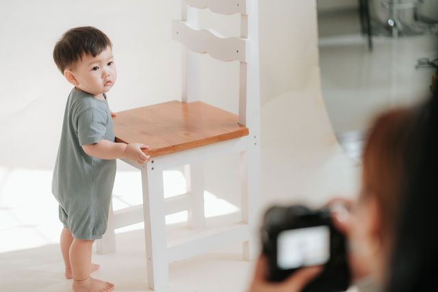 A toddler in a gray romper stands by a wooden chair during a professional photo session.