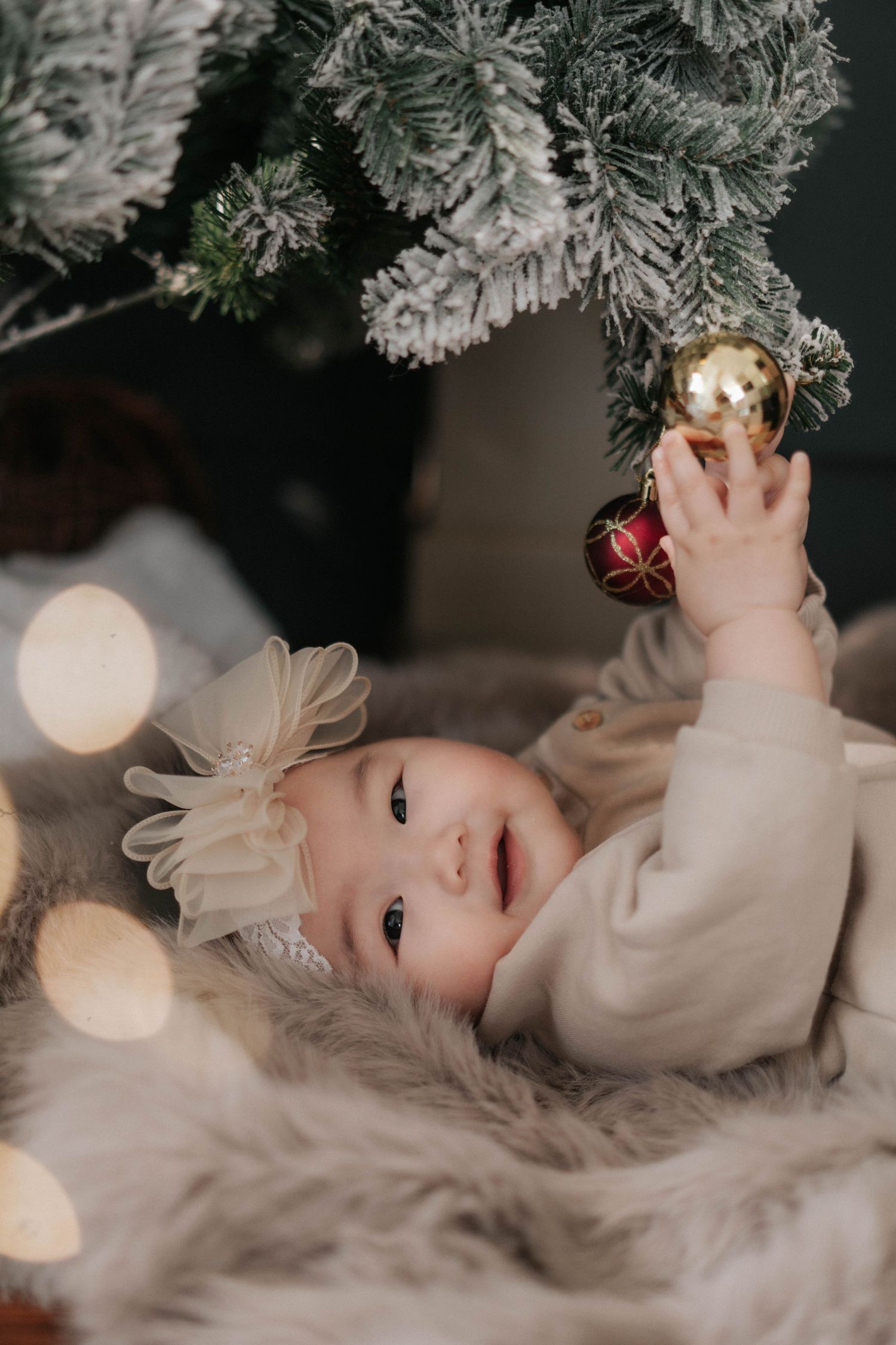 Adorable baby girl in cream outfit reaching for gold Christmas ornament under snow-flocked tree on fluffy fur rug.