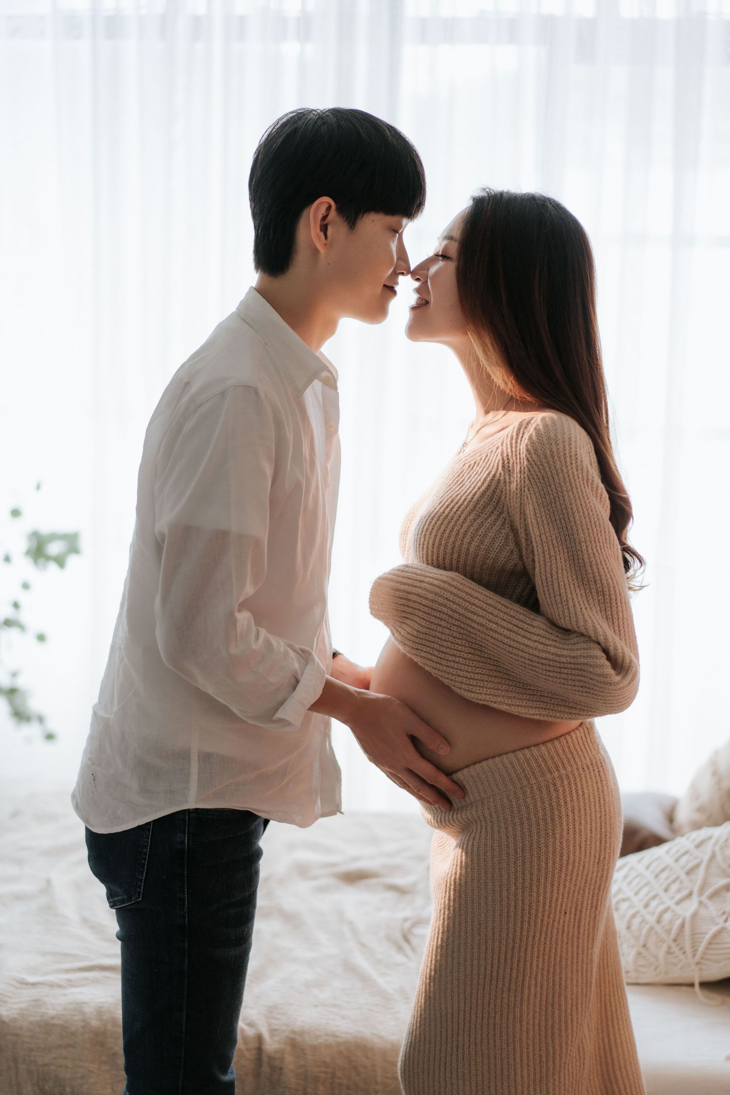A couple shares an intimate moment during a maternity photoshoot in a bright, airy bedroom setting.一對夫婦在明亮通風的臥室拍攝孕婦照，享受著親密的時刻。
