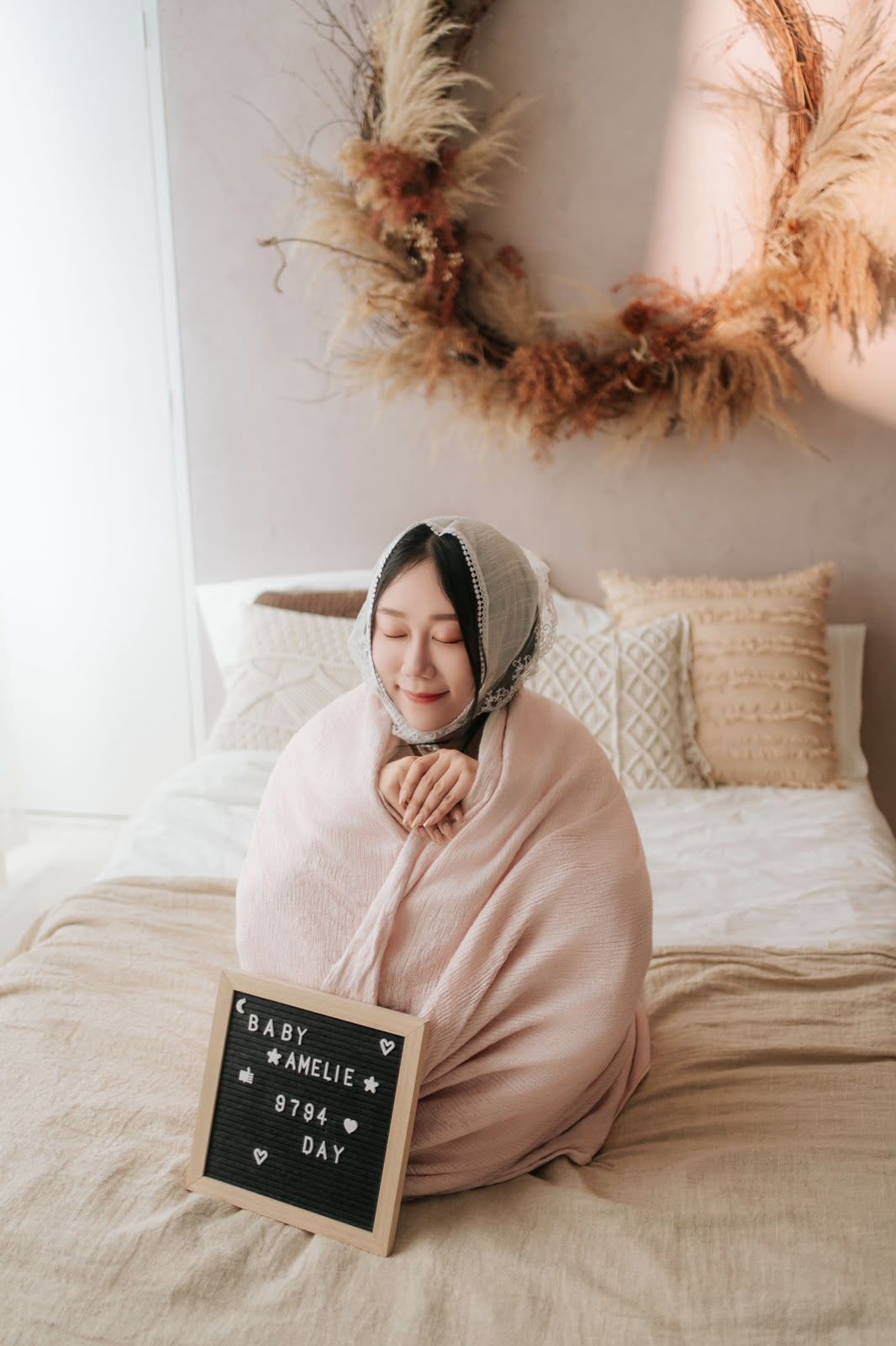 Pregnant woman wrapped in pink blanket sitting on bed with Baby Amelie letter board sign, boho nursery decor.