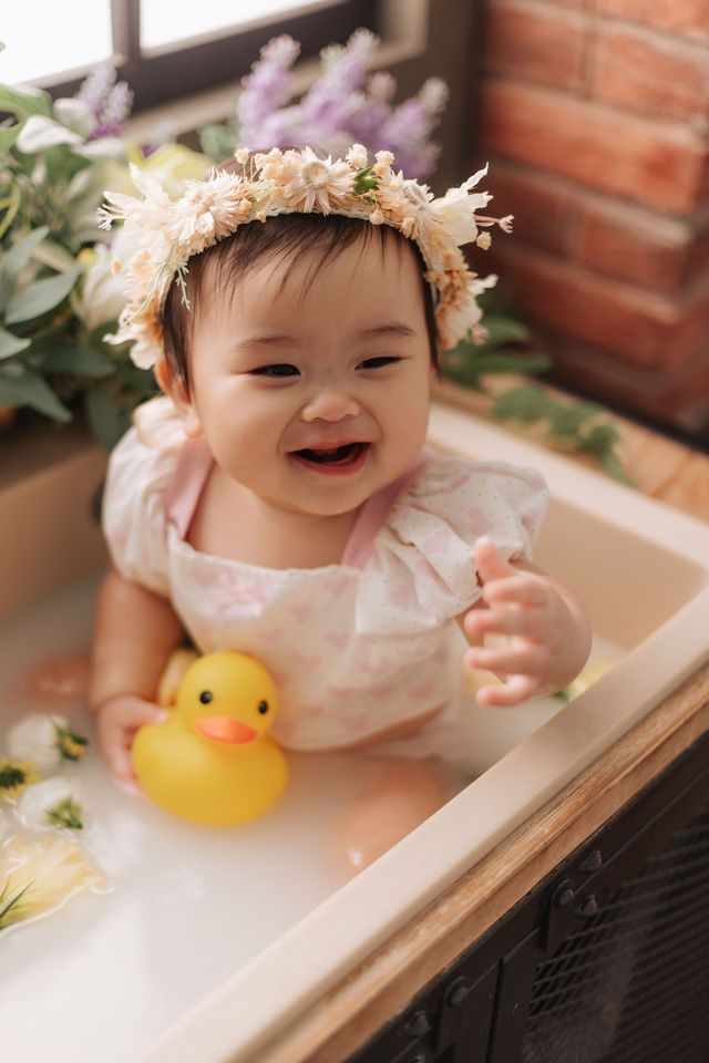 Smiling baby girl wearing floral crown sits in milk bath with yellow rubber duck, surrounded by flowers.