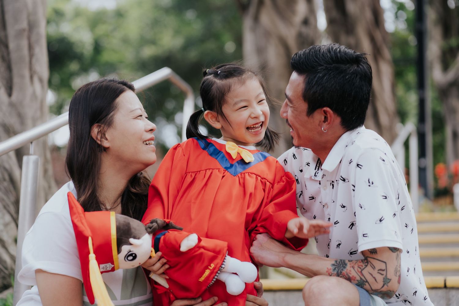 Happy family with parents smiling at laughing girl in red dress holding stuffed animal outdoors.幸福的一家人，父母微笑地看著穿著紅色洋裝、抱著毛絨玩具的小女孩在戶外開心地笑著。