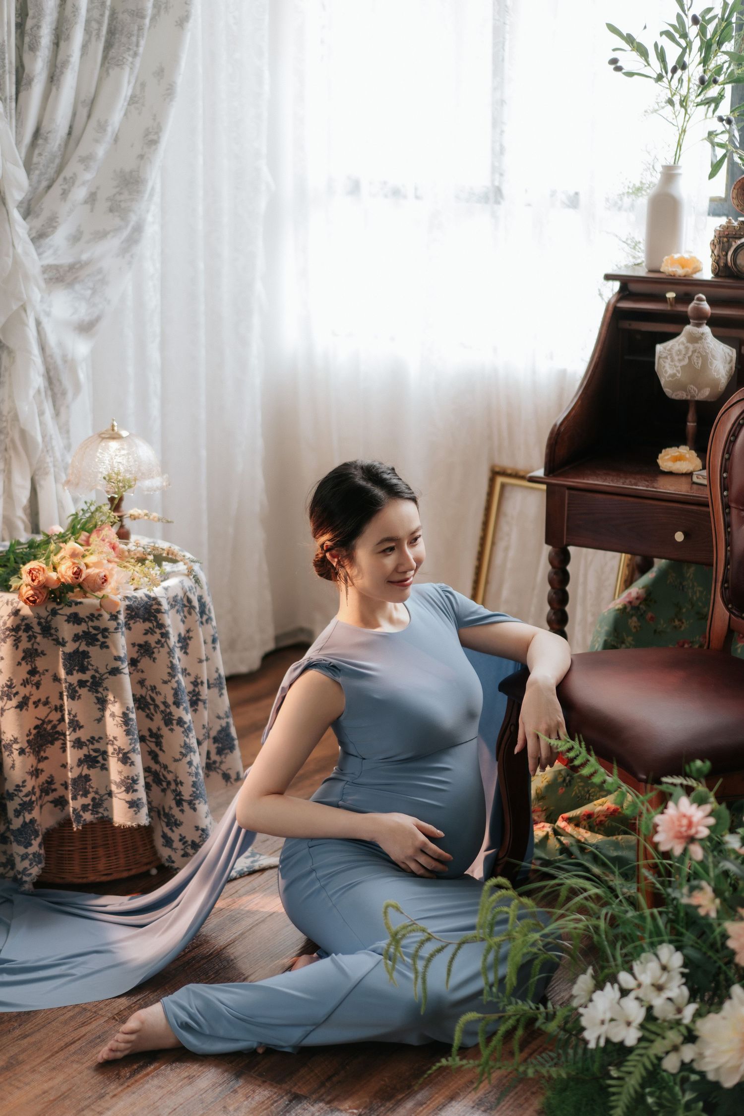Pregnant woman in blue dress reclines on vintage sofa surrounded by elegant floral arrangements and soft curtains.