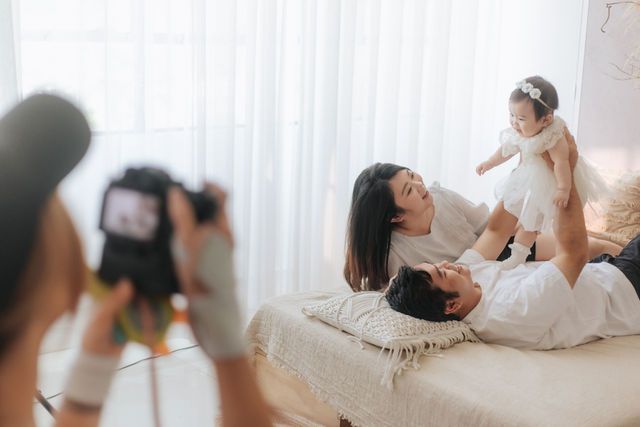 Photographer capturing a joyful family photo session with parents playing with baby on a bed near white curtains.
