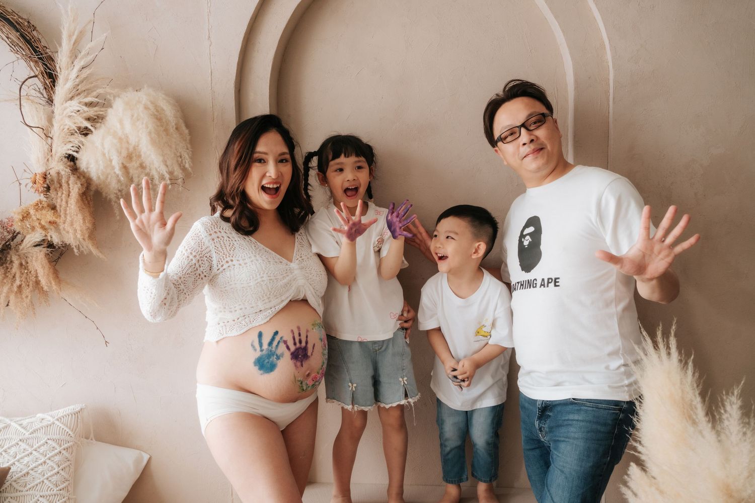 Pregnant woman with colorful handprints on belly poses with husband and two children in maternity photo shoot.一位孕婦的肚子上印著色彩鮮豔的手印，她與丈夫和兩個孩子一起
