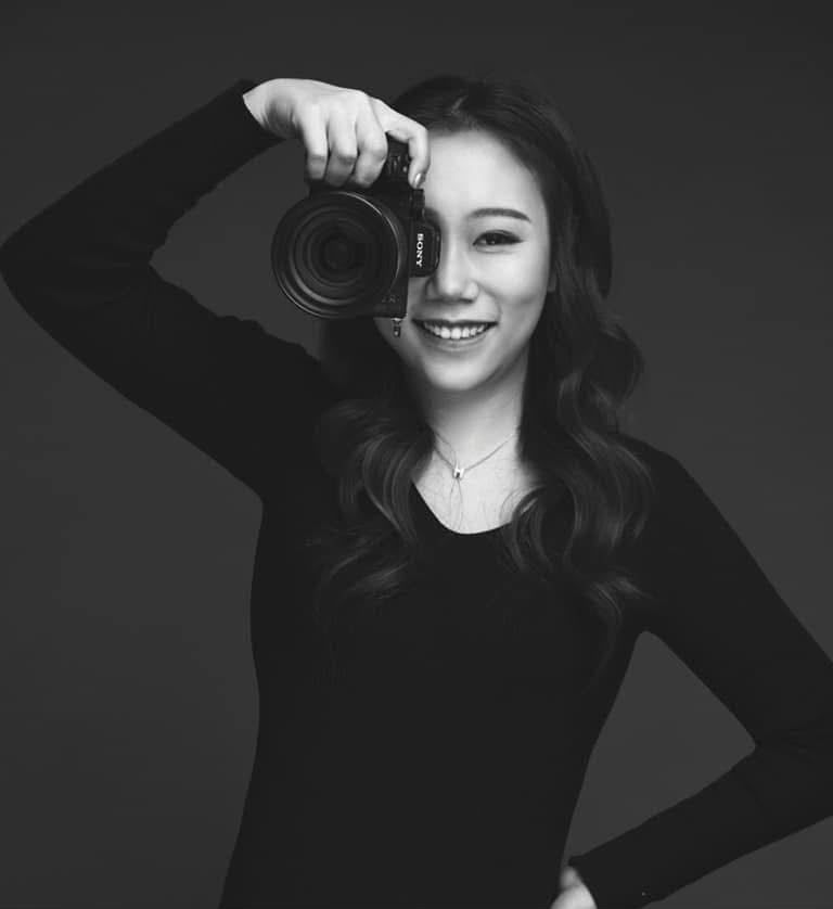 Black and white portrait of a smiling young woman holding a camera up to her eye in a studio setting.