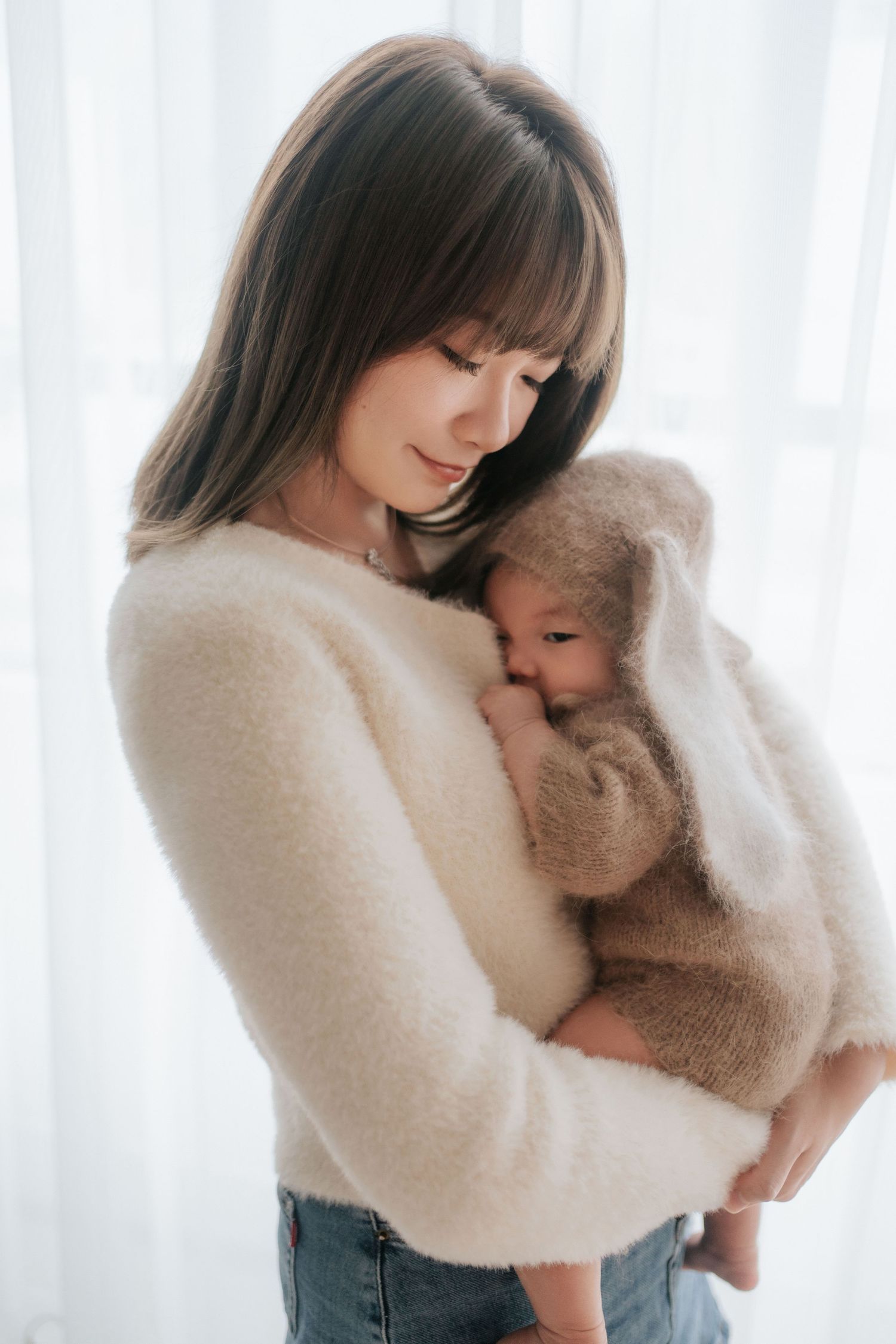 A person in a white fuzzy sweater hugs a large teddy bear against a bright window backdrop.一個穿著白色毛衣的人抱著一隻大泰迪熊，背景是明亮的窗戶。