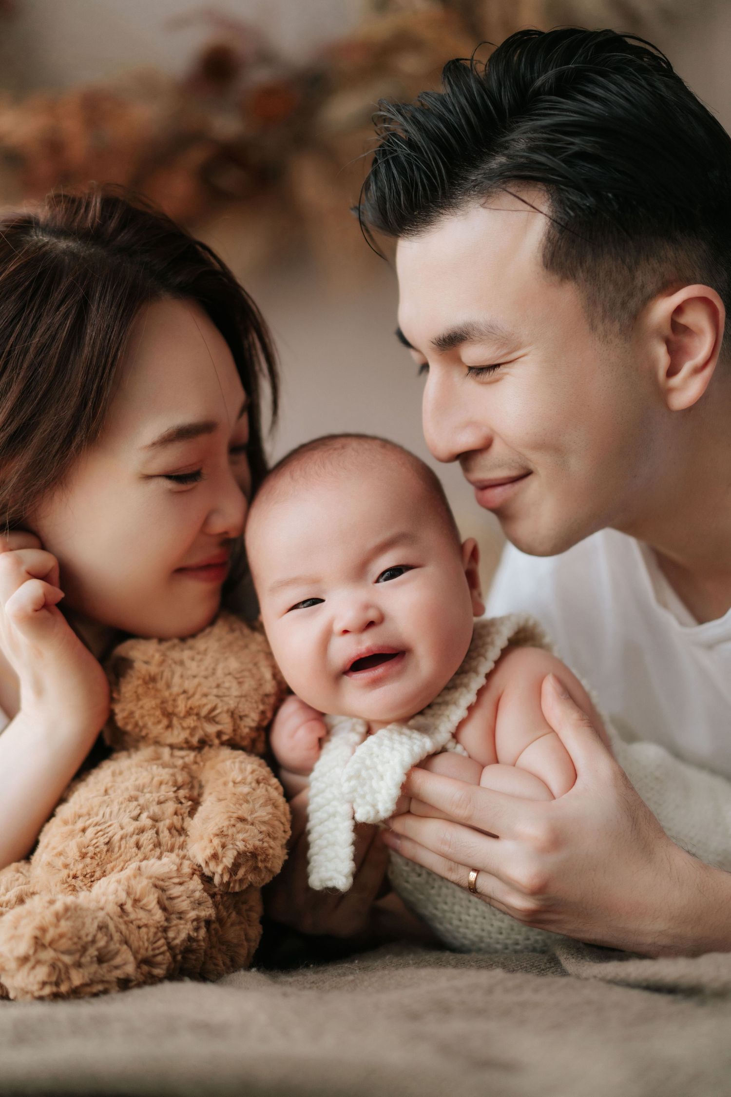 A tender family moment shows parents lovingly embracing their baby while lying down with a plush teddy bear.溫馨的家庭畫面中，父母抱著寶寶，身邊放著毛絨泰迪熊