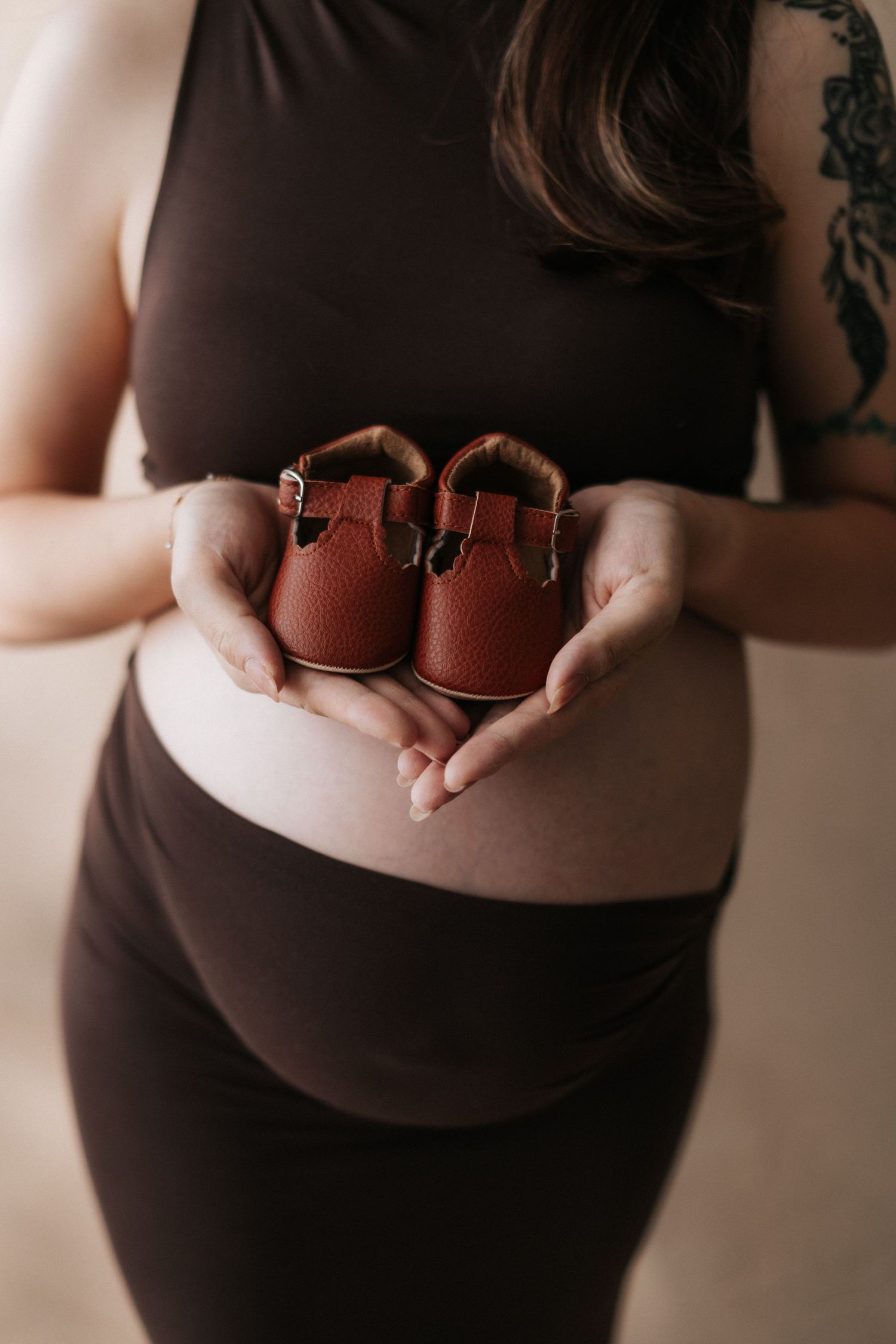 A pregnant woman in black clothing holds tiny brown leather baby shoes against her belly in a tender maternity photo.在一張溫馨的孕婦照中，一位身穿黑色衣服的孕婦
