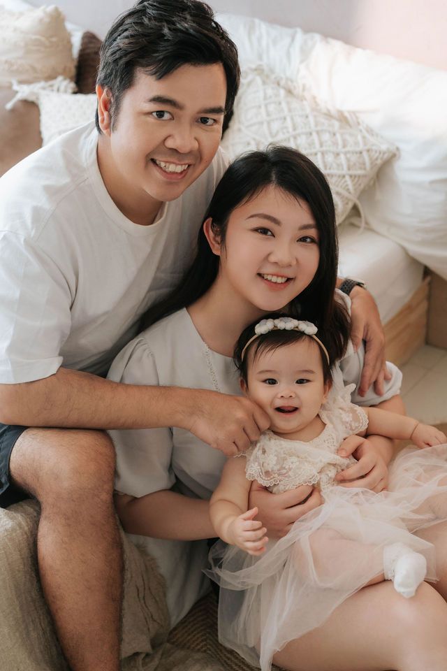 Happy Asian family with parents smiling and holding their baby girl in a white dress on a cozy bedroom floor.