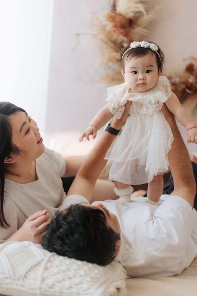 Asian parents lovingly lift their baby girl in a white tulle dress during a cozy family photo session.