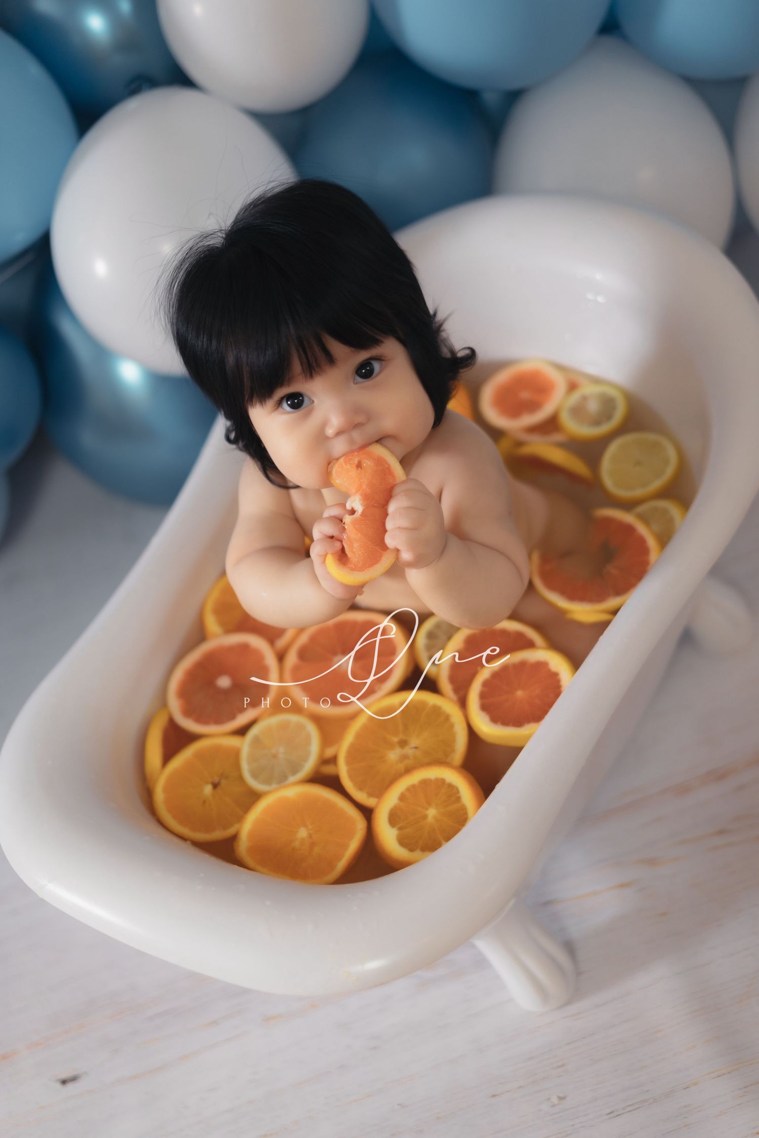 Baby sitting in a small white bathtub filled with orange slices, surrounded by blue and white balloons.嬰兒坐在裝滿橘子片的白色小浴缸裡，周圍環繞著藍色和白色的氣球。