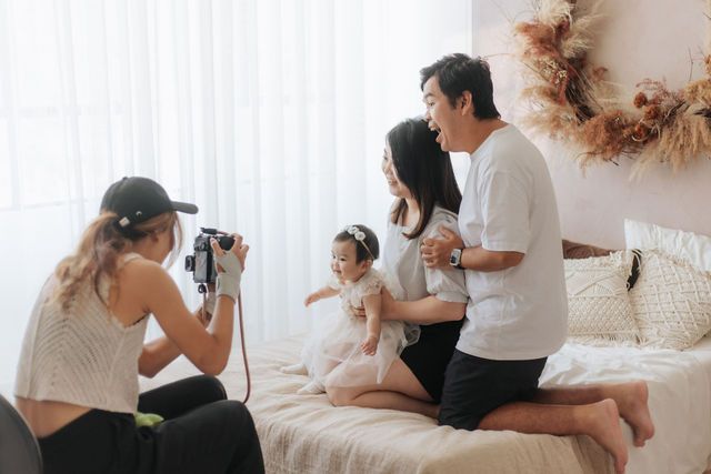 Photographer capturing Asian family portrait with toddler on bed in bright white room.