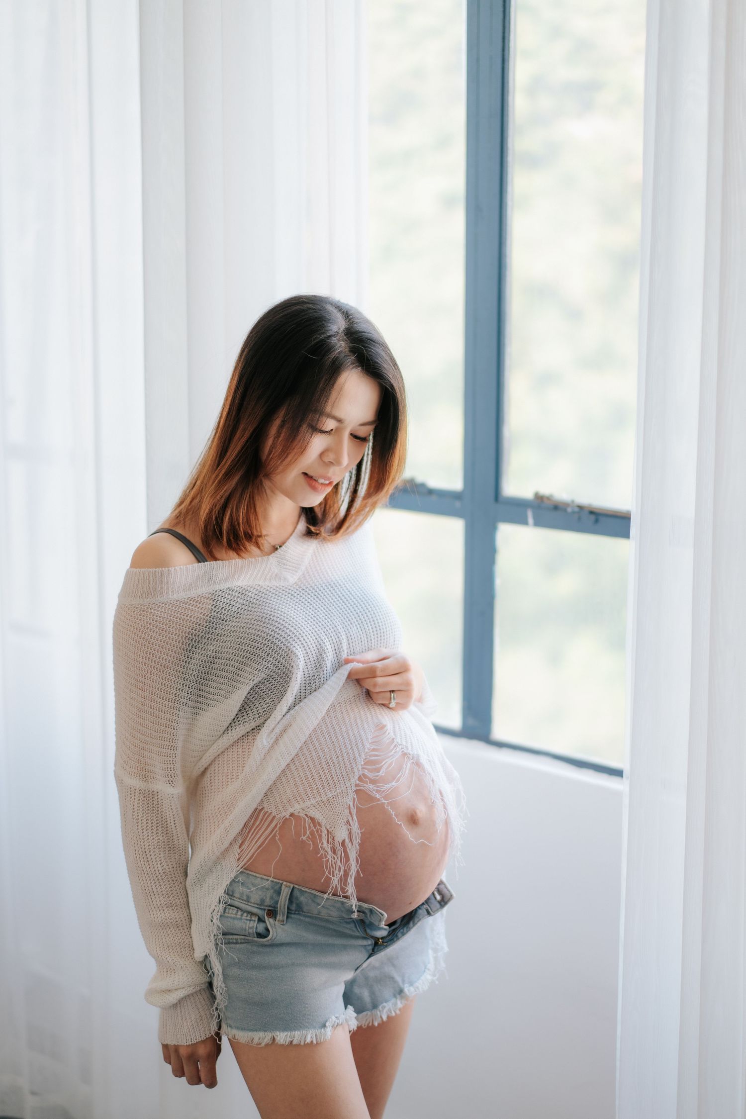 Expectant mother in white sweater and denim shorts stands by window with natural light, looking down at her pregnant belly.