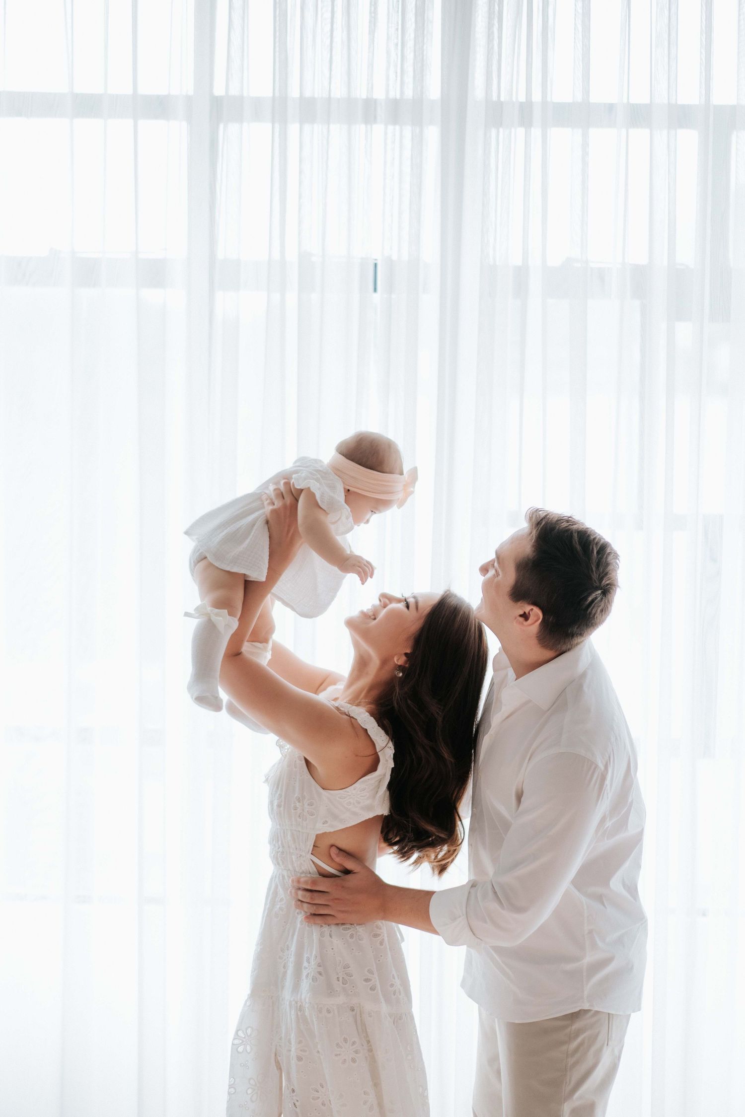 Happy family in white outfits with mother lifting smiling baby while father embraces them by bright window.幸福的一家人穿著白色服裝，母親抱著笑容燦爛的嬰兒，父親在明亮的窗邊