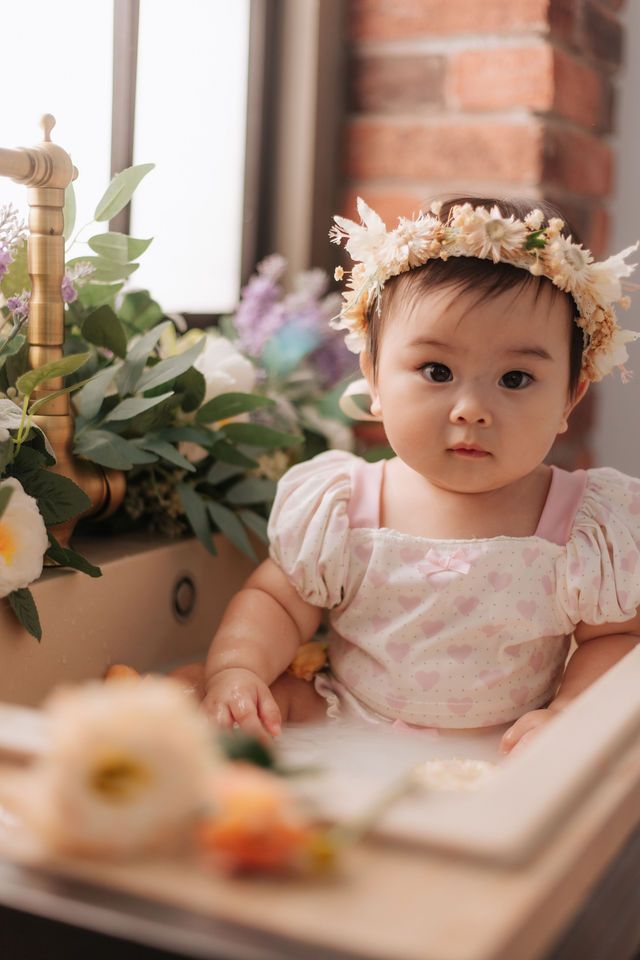 Sweet Asian baby girl in pink floral dress and flower crown sits in a decorative sink with blooms.
