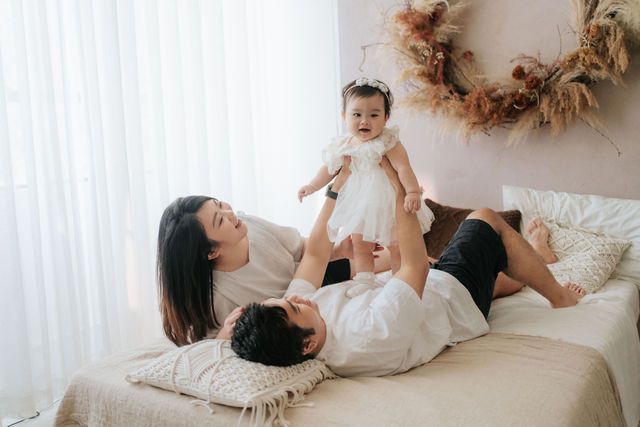 Happy young family playing with baby girl in white dress on cozy bed in bright modern bedroom.