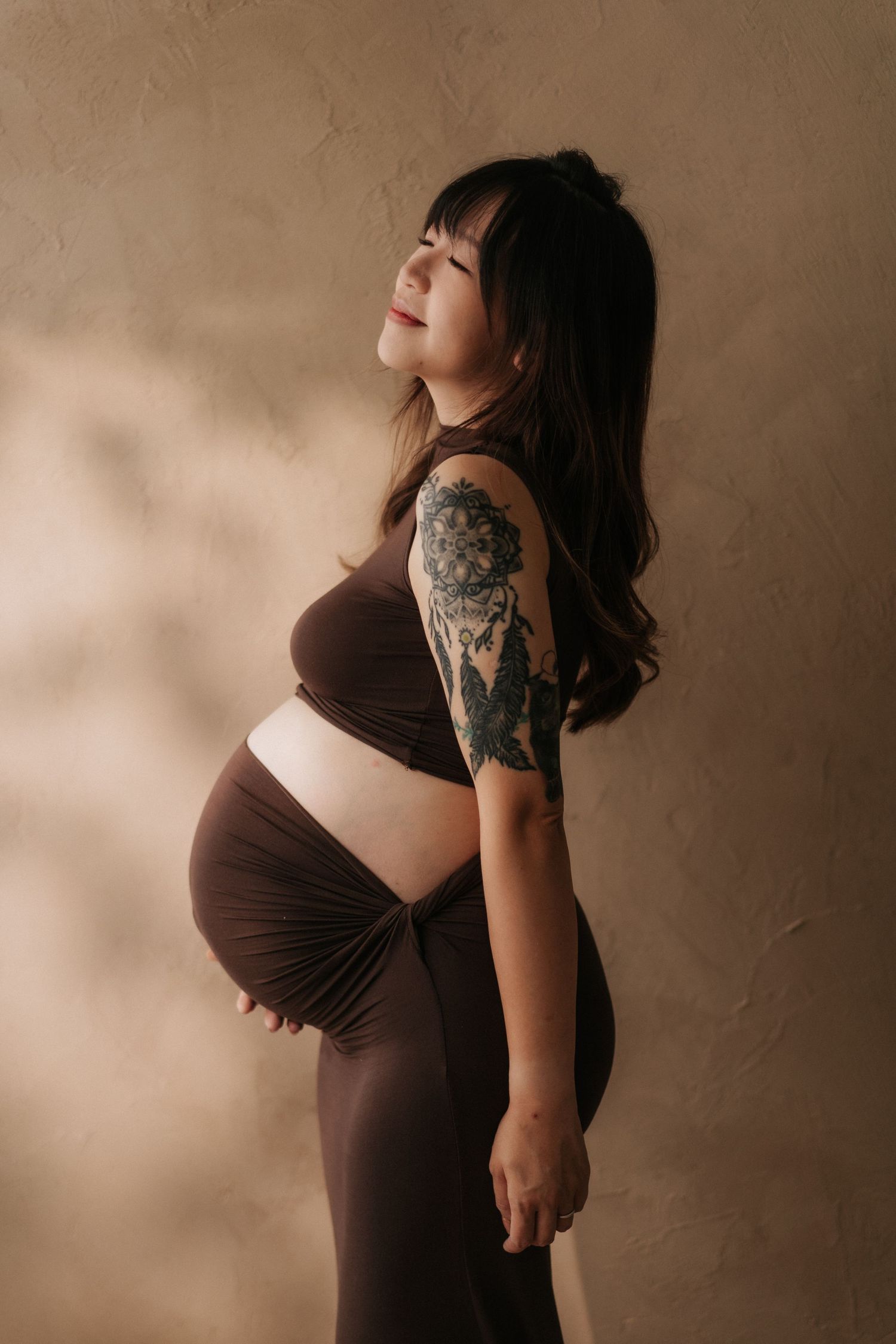 Pregnant woman with tattoo on arm posing sideways in brown crop top and skirt against beige textured wall.一位手臂上有刺青的孕婦穿著棕色露臍上衣和裙子，側身倚靠在米色紋理牆上