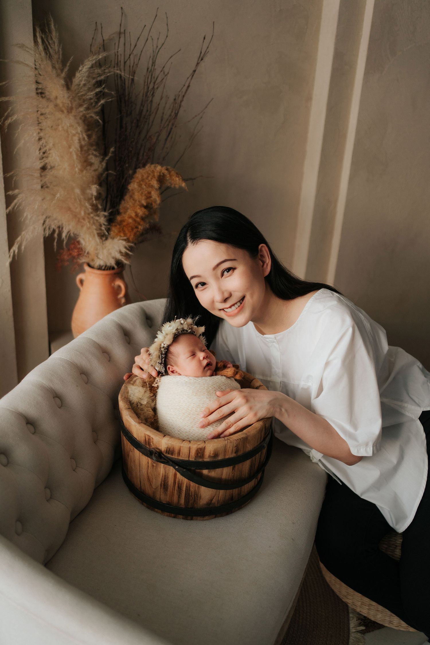 Smiling young woman posing with sleeping newborn baby wrapped in white cloth inside a wooden barrel prop.一位微笑的年輕女子抱著裹在白布里的熟睡新生兒，嬰兒被放置在木桶道具中。