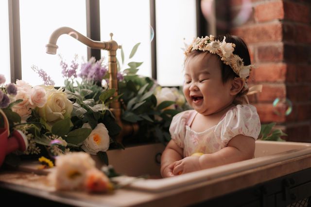 A joyful baby girl wearing a floral crown laughs while sitting in a vintage sink surrounded by beautiful flowers.