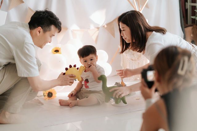 Family playing with dinosaur toys with baby on white blanket in cozy teepee setting with soft lighting.