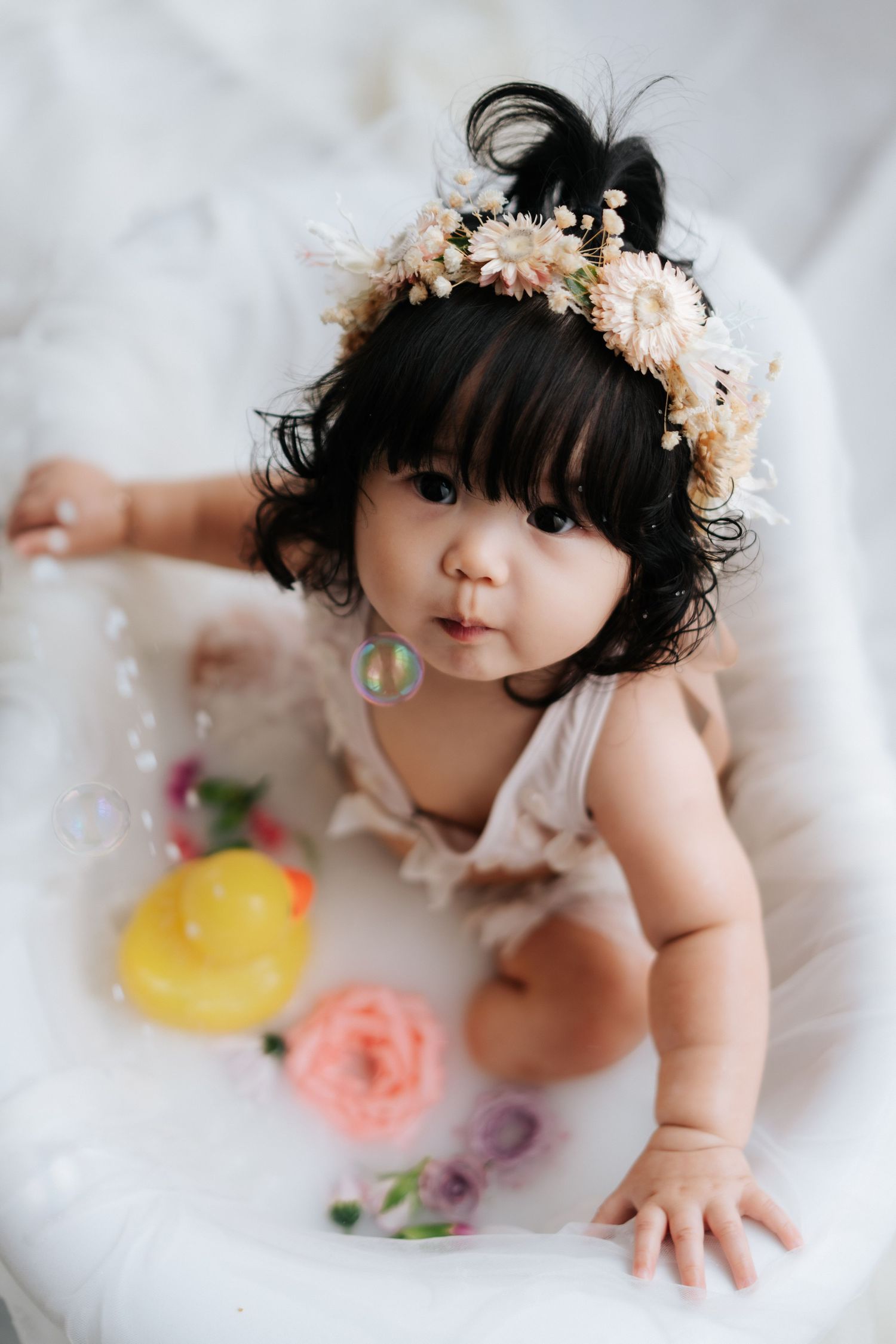 Baby in a floral crown plays with colorful bath toys in a milk bath photoshoot.戴著花冠的寶寶在牛奶浴中玩耍色彩繽紛的洗澡玩具，並拍攝了一組照片。